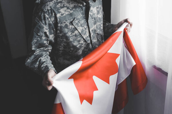 partial view of man in military uniform holding canada national flag while standing by window