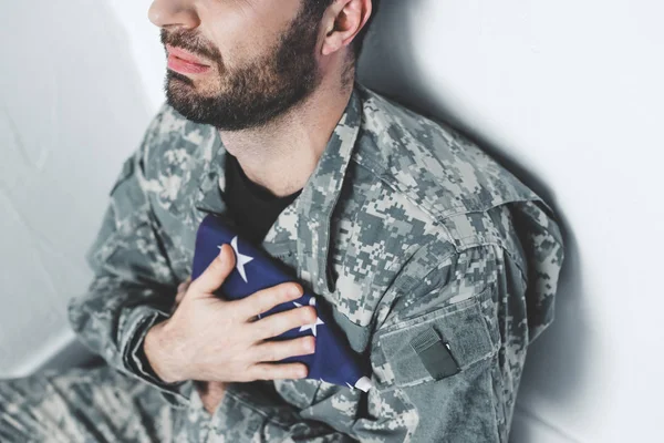 partial view of depressed military man sitting by white wall and holding usa military man near heart