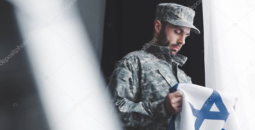 selective focus of pensive military man in uniform holding israel national flag while standing by window