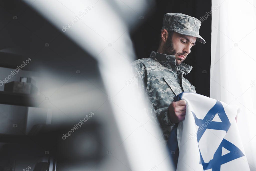 selective focus of pensive bearded man in military uniform holding israel national flag while standing by window