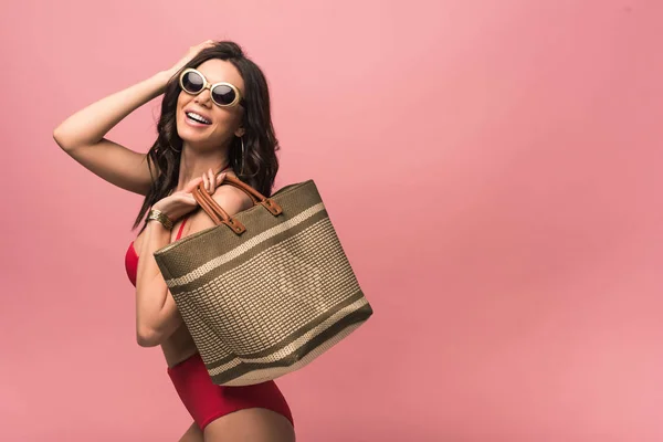 微笑有吸引力的女人在泳衣和太阳镜拿着海滩袋孤立在粉红色 — 图库照片