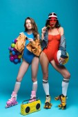 full length view of two stylish girls in roller skates eating watermelon lollipop on blue