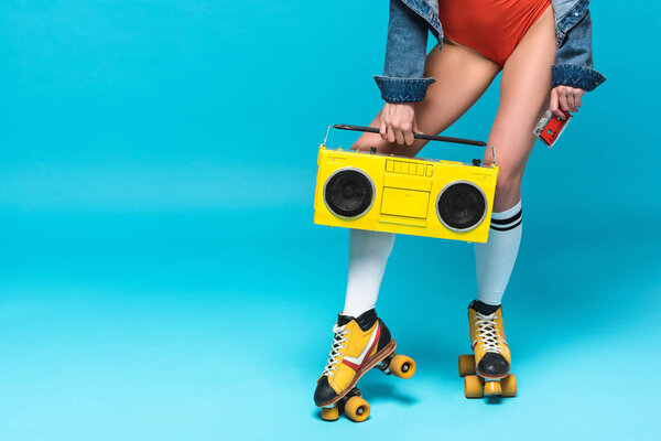 cropped view of woman in swimsuit and roller skates holding boombox and cassette tape on blue
