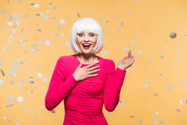surprised girl in red dress and white wig posing with holiday confetti, isolated on yellow