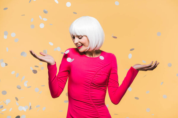 cheerful pretty girl in red dress and white wig posing with holiday confetti, isolated on yellow