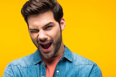 excited handsome man winking and looking at camera isolated on yellow clipart