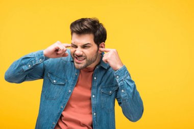 dissatisfied handsome man plugging ears with fingers isolated on yellow clipart
