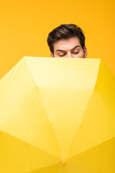 handsome man covering face with umbrella and looking down isolated on yellow