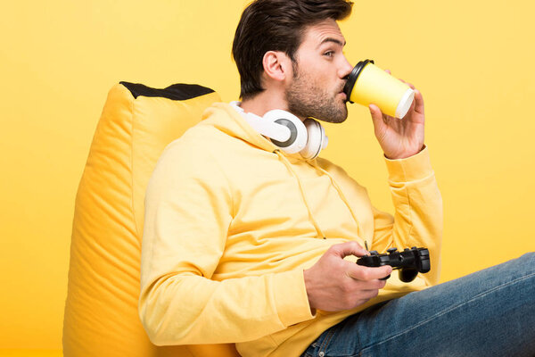 KYIV, UKRAINE - APRIL 12: man drinking coffee to go on bean bag chair and playing Video Game isolated on yellow