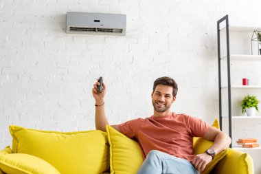handsome man looking at camera while sitting under air conditioner and holding remote control clipart