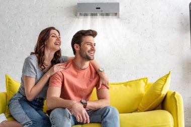 cheerful couple sitting on yellow sofa under air conditioner at home clipart