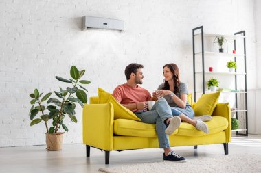 happy young couple talking and holding cups while sitting on yellow sofa under air conditioner at home clipart