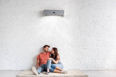 happy young couple hugging while sitting on floor near white wall under air conditioner clipart