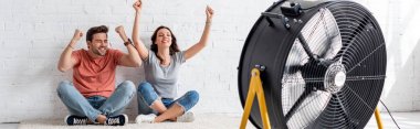 panoramic shot of excited man and woman sitting on floor with raised hands in front of blowing electric fan clipart