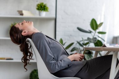exhausted businesswoman sitting on chair with closed eyes and holding glass of water while suffering from heat in office clipart