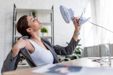 selective focus of beautiful businesswoman suffering from heat and holding electric fan clipart
