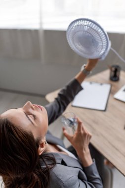 overhead view of pretty businesswoman holding blowing electric fan and glass of water while suffering from heat in office clipart