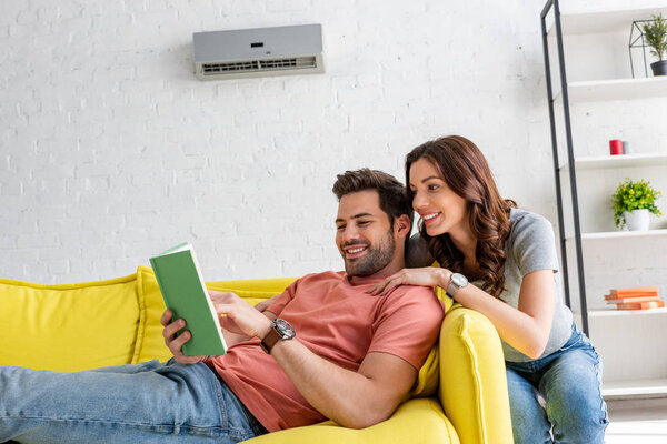handsome man reading book while lying on sofa under air conditioner near smiling woman