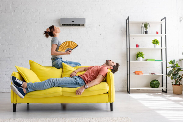 измученный мужчина лежит на желтом диване под кондиционером рядом с женщиной с ручным вентилятором
