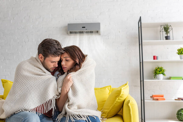 frozen couple warming under blanket while sitting on sofa under air conditioner at home