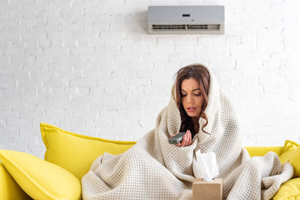 frozen sick woman with remote control warming under fleece blanket while sitting on sofa under air conditioner at home