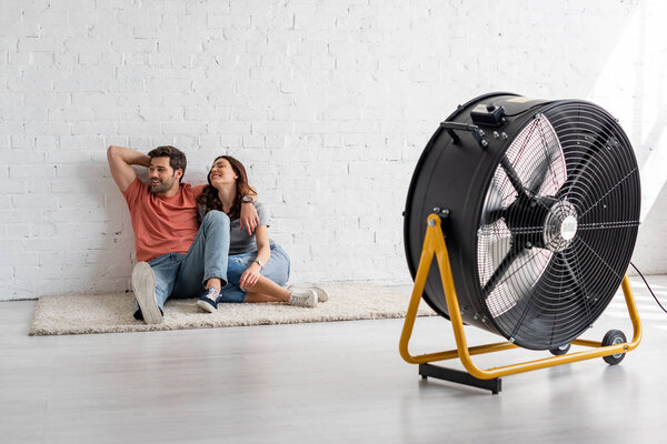 young smiling couple sitting on floor by white wall in front of blowing electric fan 
