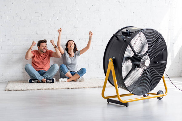 excited man and woman sitting on floor by white wall in front of blowing electric fan