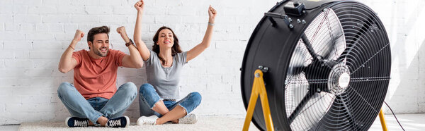 panoramic shot of excited man and woman sitting on floor with raised hands in front of blowing electric fan