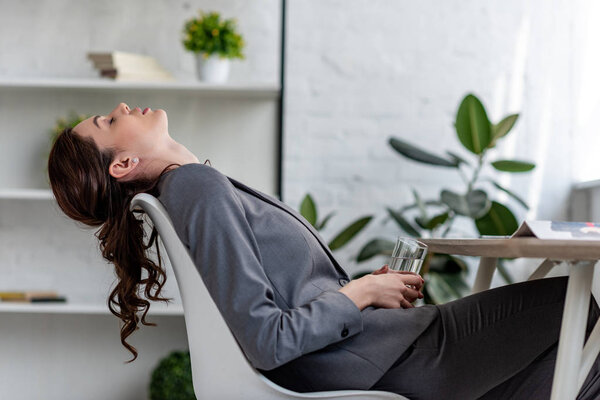 exhausted businesswoman sitting on chair with closed eyes and holding glass of water while suffering from heat in office