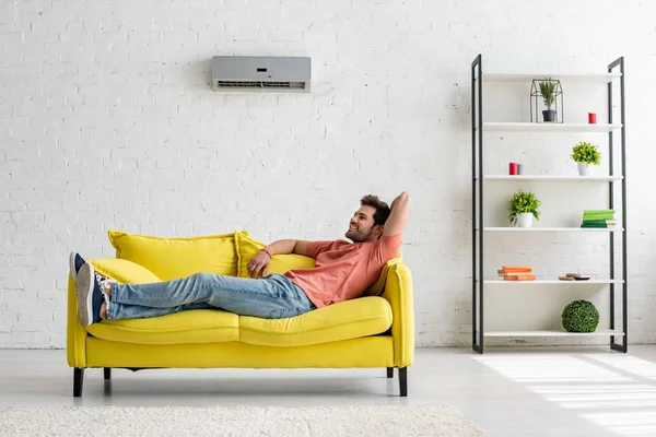 Handsome smiling man lying on yellow sofa under air conditioner at home — Stock Image Handsome Smiling Man Lying Yellow Sofa Air Conditioner Home Stock Photo