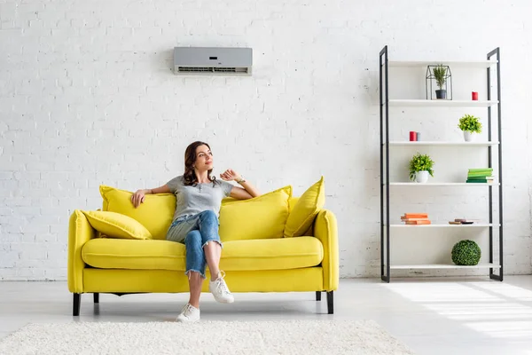 Beautiful young woman sitting on yellow sofa under air conditioner in spacious apartment — Stock Image Beautiful Young Woman Sitting Yellow Sofa Air Conditioner Spacious Apartment Stock Image