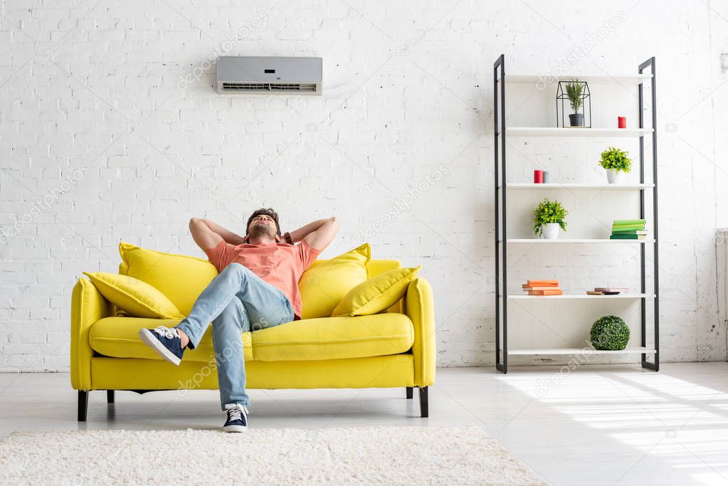 young man sitting on yellow sofa under air conditioner in spacious apartment