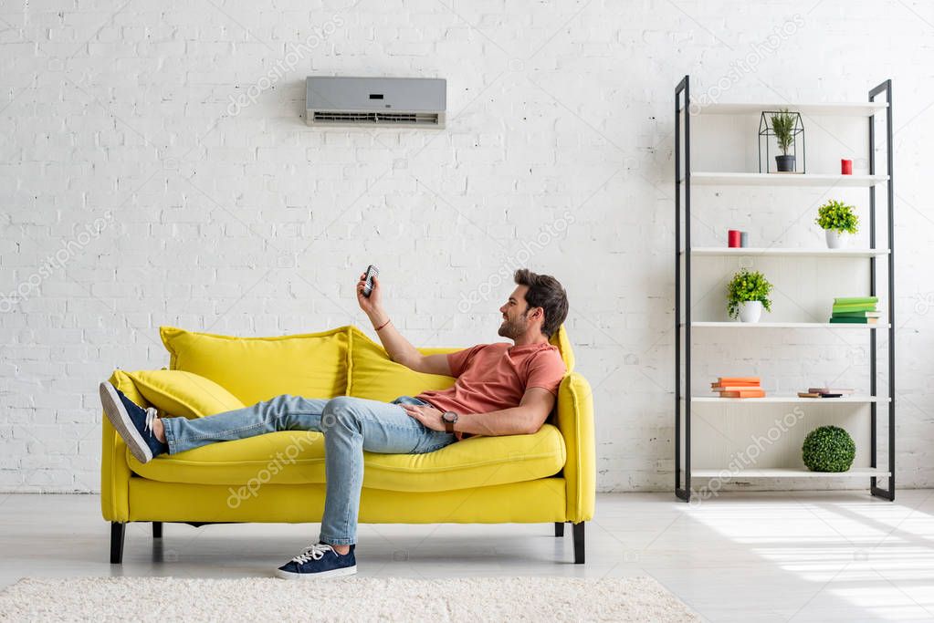 handsome young man lying on yellow sofa under air conditioner and holding remote control