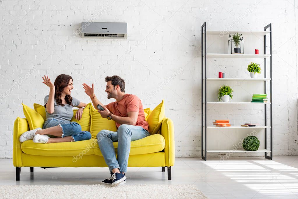 cheerful man and woman talking while sitting on yellow sofa under air conditioner