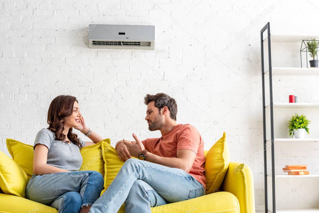 cheerful man and woman talking while sitting on yellow sofa under air conditioner at home