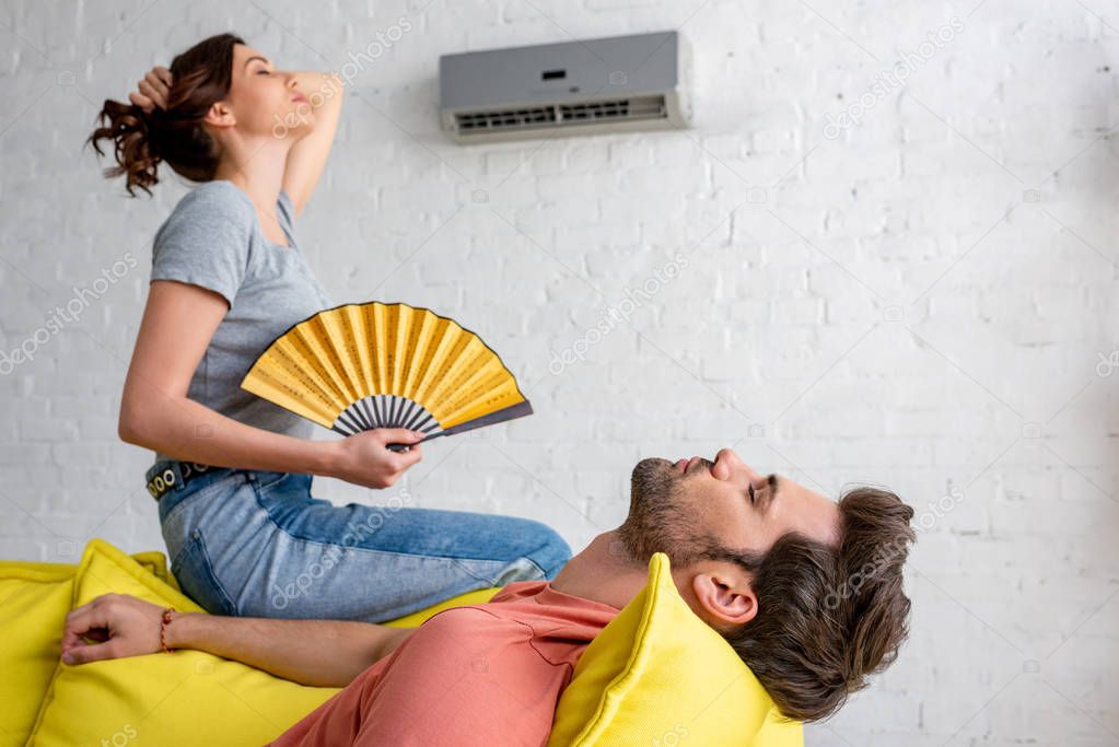 young woman with hand fan sitting on yellow sofa under air conditioner near lying man 