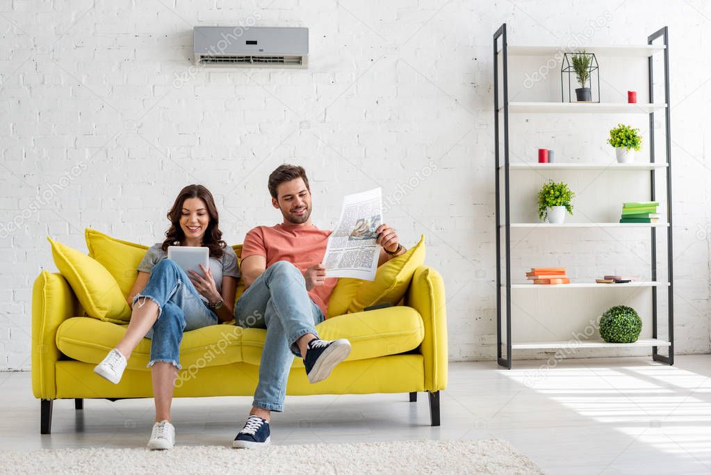 pretty woman using digital table and handsome man reading newspaper while sitting on yellow sofa under air conditioner at home