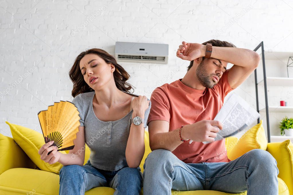 pretty woman with hand fan and handsome man with newspaper suffering from heat at home