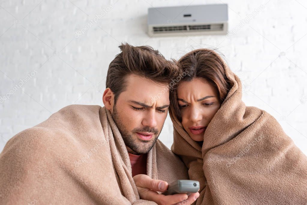 frozen man and woman warming under blankets while sitting under air conditioner with remote control