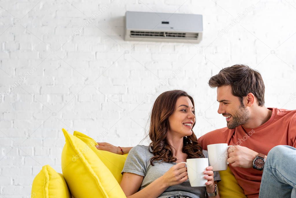 happy couple with cups sitting on sofa under air conditioner at home