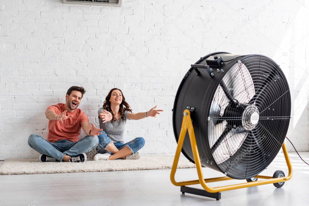 excited man and woman sitting on floor with outstretched hands in front of electric ventilator