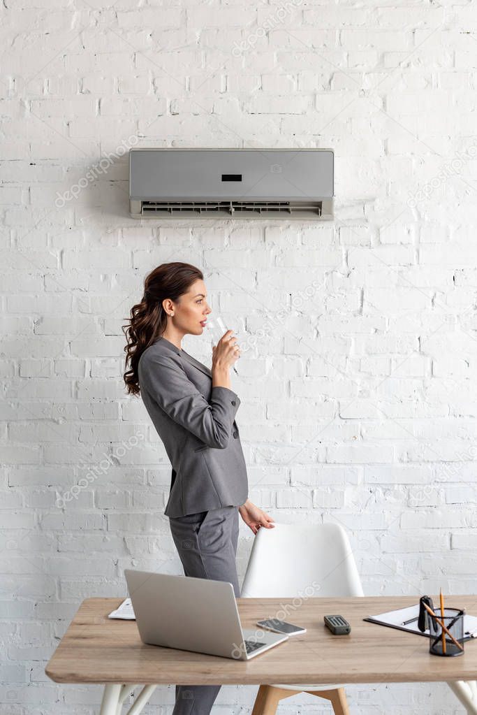 attractive businesswoman drinking water while standing near workplace under air conditioner