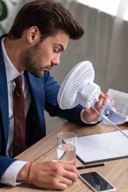 exhausted businessman sitting in front of blowing electric fan while suffering from heat in office clipart