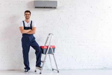 smiling repairman standing under air conditioner near stepladder and toolbox and looking at camera clipart