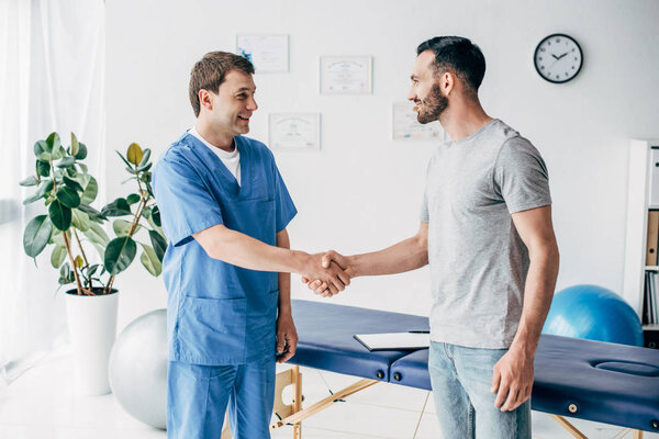 Smiling patient and doctor shaking hands near couch in massage cabinet at clinic