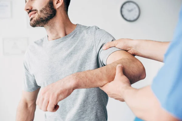 痛みに苦しむ患者の選択的焦点とクリニックのマッサージキャビネットで患者の腕を検査する医師 — ストック写真