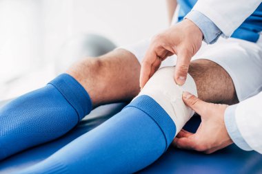 partial view of Physiotherapist putting elastic bandage on leg of football player clipart
