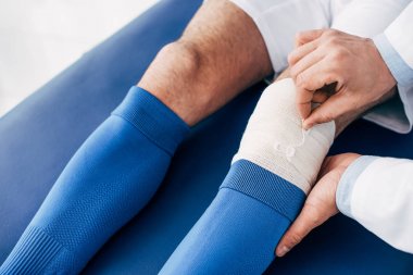 cropped view of Physiotherapist putting elastic bandage on leg of football player clipart