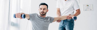 panoramic shot of chiropractor stretching arm of handsome patient with dumbbells in hospital clipart