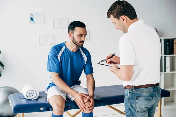 Fisioterapeuta Escribiendo Diagnóstico Mientras Futbolista Sentado Mesa Masaje Hospital — Foto de Stock